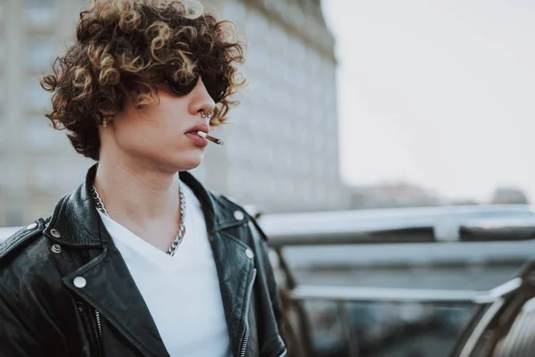 Curly rocker in sunglasses standing alone and smoking