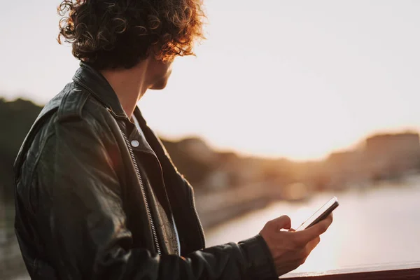 Rocker rizado mirando el atardecer y sosteniendo el teléfono inteligente — Foto de Stock