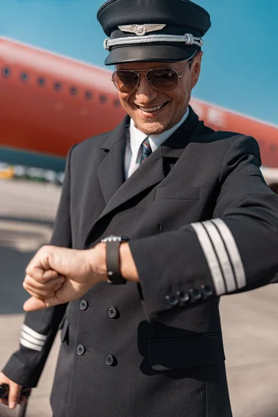 Веселый кавказский пилот в форме смотрит на часы в аэропорту — стоковое фото