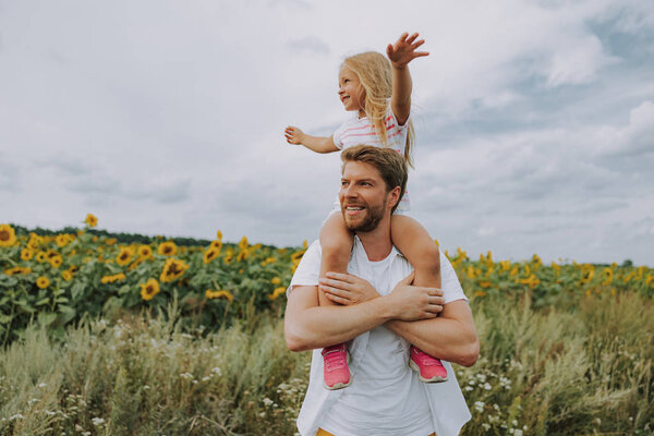 Счастливая веселая дочь сидит на своих улыбающихся папах плечах на улице
