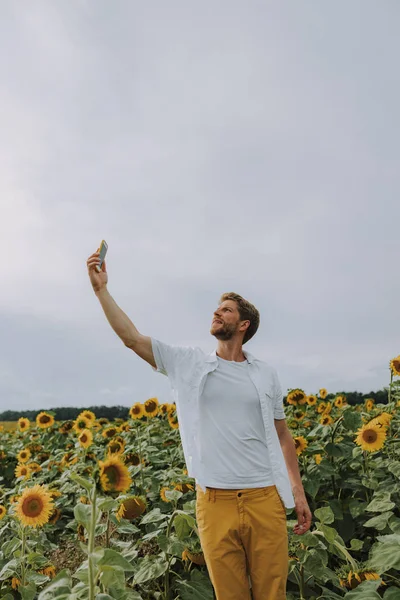 Schattige Kaukasische kerel maakt selfie met telefoon in zonnebloem veld in de zomer — Stockfoto