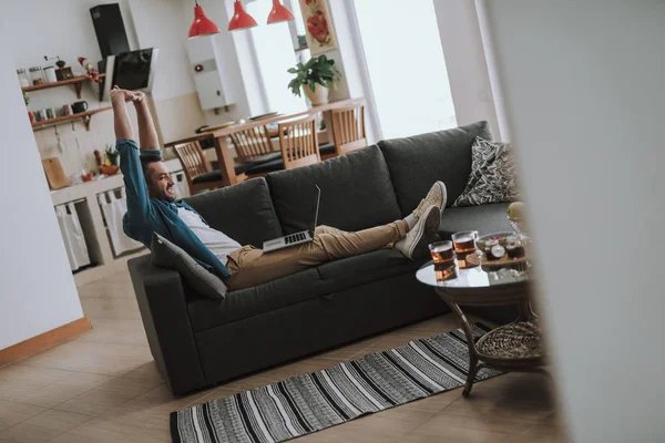Pemuda tampan dengan laptop merentangkan tangan sambil beristirahat di sofa di rumah — Stok Foto
