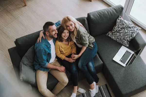 Happy parents hugging their daughter while sitting on couch with laptop — Stock Photo, Image