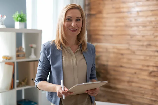 Waist up of smiling female psychologist holding clipboard and pen in arms