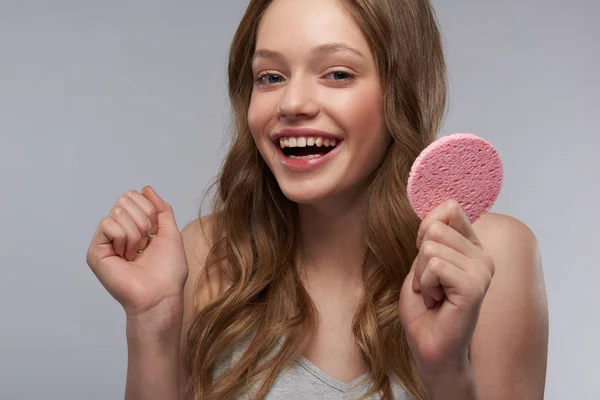 Menina adolescente alegre com esponja cosmética em pé contra fundo cinza-branco — Fotografia de Stock