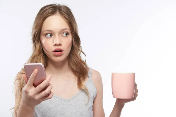 Verbaasd tiener meisje dat roze mobiele telefoon en kopje koffie vasthoudt — Stockfoto