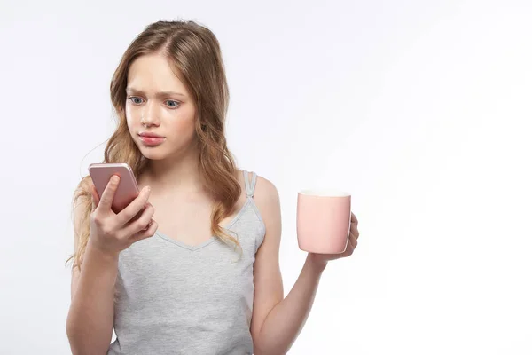 Geschokt tiener meisje houden kopje koffie tijdens het gebruik van mobiele telefoon — Stockfoto