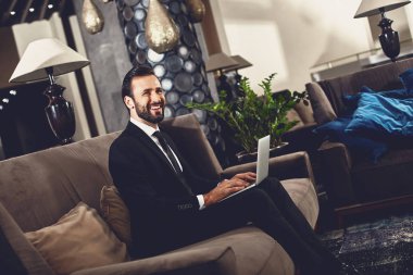 Sakallı genç adam modern cihazı ile kanepede otururken gülümseyen