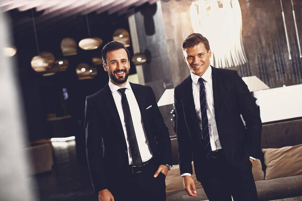 Два стильных мужчины улыбаются, стоя в холле вместе
