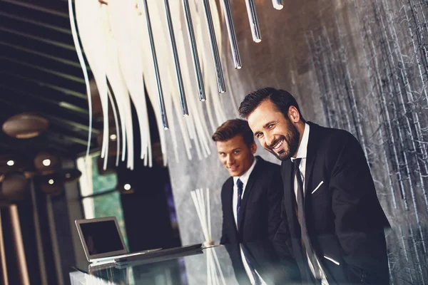 Homens do hotel alegres sentados na recepção e sorrindo enquanto olham para você — Fotografia de Stock