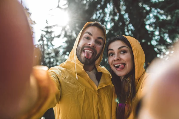 Casal de viajantes fazendo selfie engraçado em capas impermeáveis — Fotografia de Stock