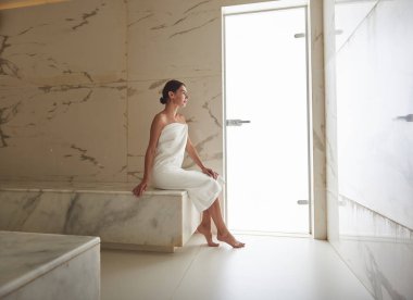 Beyaz havlu giymiş, sırtı dimdik oturan zayıf bir kadın.