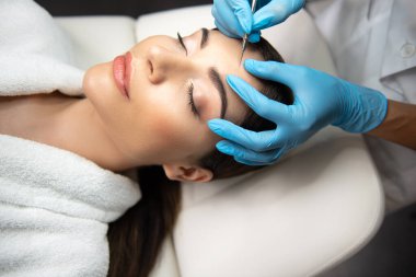 Güzellik uzmanı, siyah başlığı bayan alnından özel çelik bir aletle çıkarıyor.