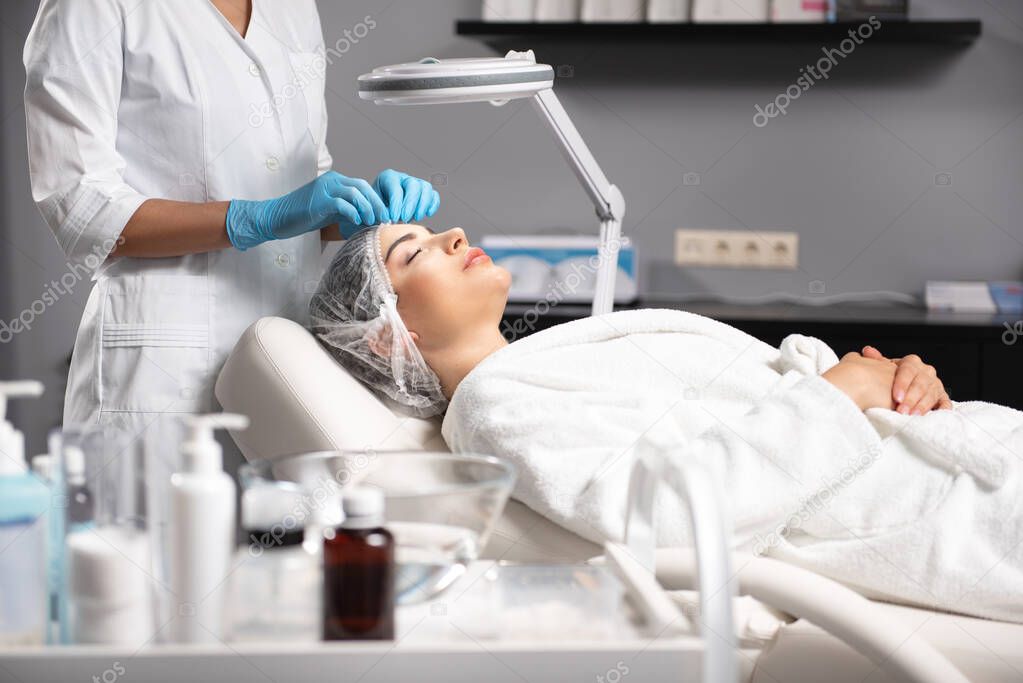 Beautician putting on disposable medical cap on lady before procedure