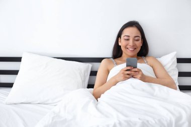 Yatak odasında akıllı telefon kullanan neşeli genç bir kadın.