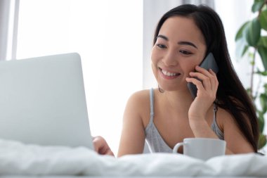 Cep telefonuyla dizüstü bilgisayarın önünde iletişim kuran mutlu kadın.