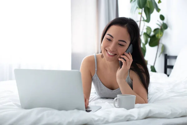 Happy woman communicating on smartphone and laptop in bed — Stock Photo, Image