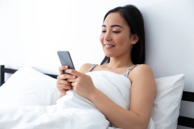 Gülümseyen kadın yatakta akıllı telefondan mesaj atıyor.