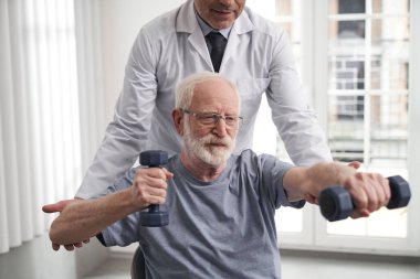 Gülümseyen yaşlı adam rehabilitasyon uzmanıyla vücut egzersizi yapıyor.