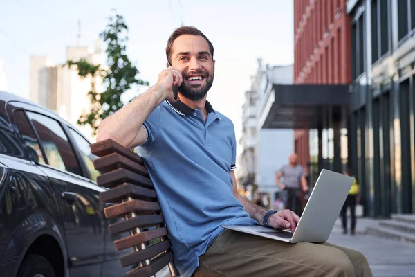 Homem feliz com notebook falando no smartphone ao ar livre — Fotografia de Stock