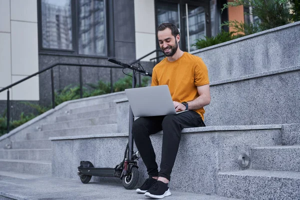 Jovem alegre com scooter usando laptop ao ar livre — Fotografia de Stock