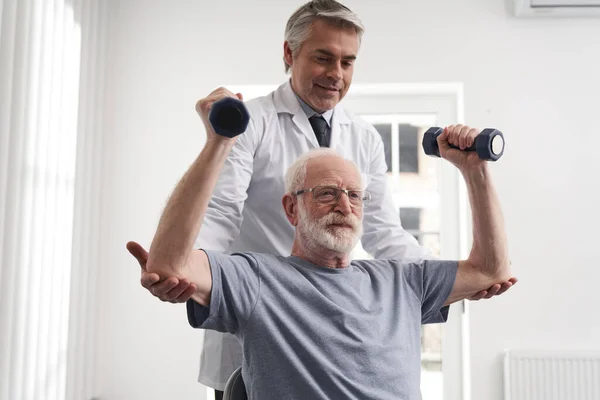 Neşeli olgun doktor yaşlı adama egzersizlerinde yardım ediyor. — Stok fotoğraf