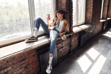 Genç adam pencere eşiğinde uzanıyor ve telefonu tutuyor.