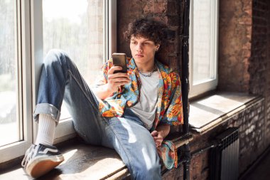 Genç adam pencere kenarında dinleniyor ve telefonu tutuyor.