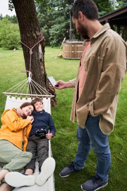 Sakallı adam, karısını ve oğlunu akıllı telefonuyla çekiyor.