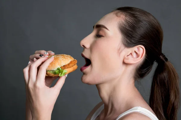 Young woman eating hamburger on gray background. Junk food and fast food concept