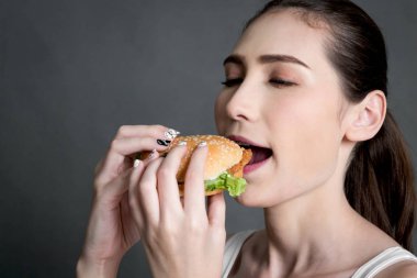Genç kadın gri arka plan üzerinde hamburger yeme. Abur cubur ve fast food kavramı
