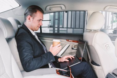 Bir arabanın arka koltuğunda oturan dizüstü bilgisayar üzerinde çalışan yakışıklı işadamı