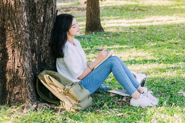 公園の芝生の上に座ってノートを書くペンを持つ女性 — ストック写真