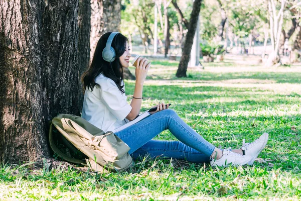 女性は 公園の草の上に座って音楽を聴くヘッドフォンで飲むコーヒーをリラックスします — ストック写真