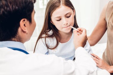 Doktor termometreyi tutuyor ve hasta küçük kızı kontrol ediyor. Ateşli küçük kız yatakta yatıyor.