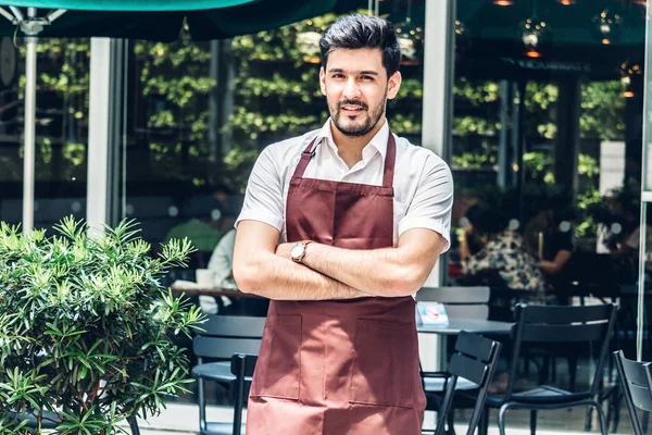Portrait of handsome small business owner smiling and standing with crossed arms outside the cafe or coffee shop.Male barista standing at cafe