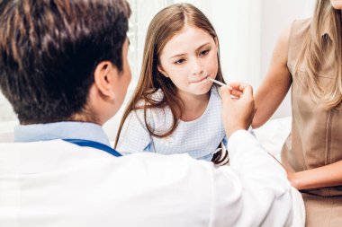 Doktor termometreyi tutuyor ve hasta küçük kızı kontrol ediyor. Ateşli küçük kız yatakta yatıyor.