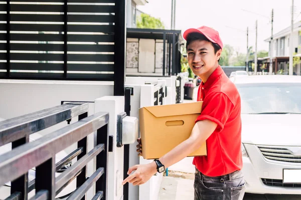 Happy delivery man in red uniform holding cardboard box standing near house of customer.courier service concept