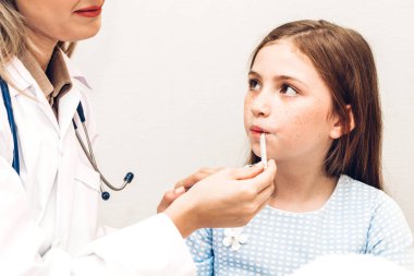 Doktor termometreyi tutuyor ve hasta küçük kızı kontrol ediyor. Ateşli küçük kız yatakta yatıyor.