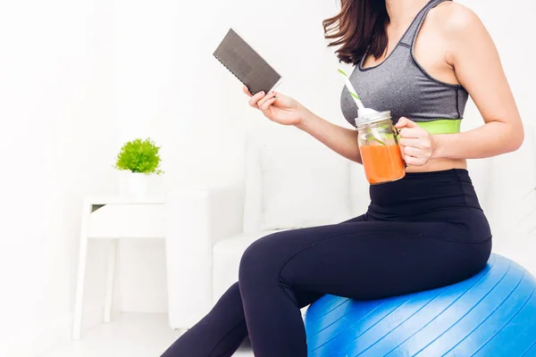 Sport woman in sportswear sitting relax reading a book and drink fresh juice after workout on blue fitball at home.Diet concept.Fitness and healthy lifestyle