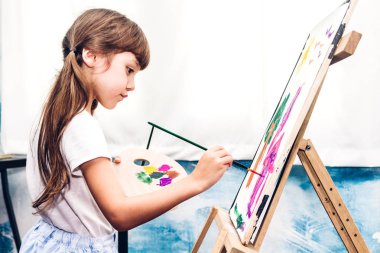 Küçük kız ressam evde renk paleti ve suluboya resimlerle tuvale resim çiziyor.