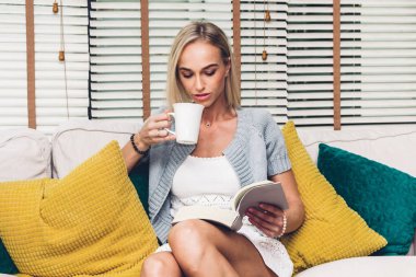 Rahatlatıcı ve bir kitap okuma ve içme kahve onun oturma odasında kanepede oturan güzel kadın