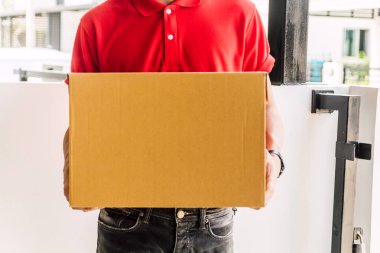 Kırmızı üniformalı mutlu teslimatçı elinde karton kutuyla müşteri evinin yanında duruyor. Kurye servisi konsepti.