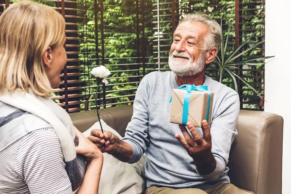 Happy smiling elder senior man giving anniversary gift box surprise to wife and talking together sitting on sofa in living room at home.Retirement couple concept