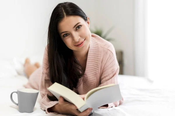 Νεαρή γυναίκα χαλαρωτικό και διαβάζοντας το βιβλίο στο κρεβάτι στο σπίτι — Φωτογραφία Αρχείου