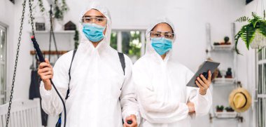 Koruyucu maske takan dezenfekte işçileri ve müşteri evindeki Coronavirüs 'ü öldürmek için beyaz takım elbise dezenfektan sprey temizlik virüsü kullanan profesyonel ekipler.