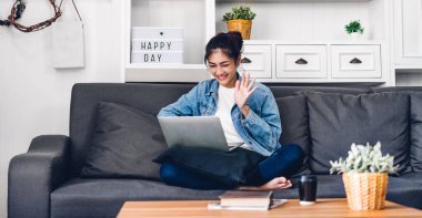 Genç gülümseyen mutlu Asyalı kadın dizüstü bilgisayarını kullanarak rahatlıyor ve evde video konferansı yapıyor. Genç yaratıcı kız merhaba diyor ve klavyede yazıyor. Ev konseptinden çalışma.