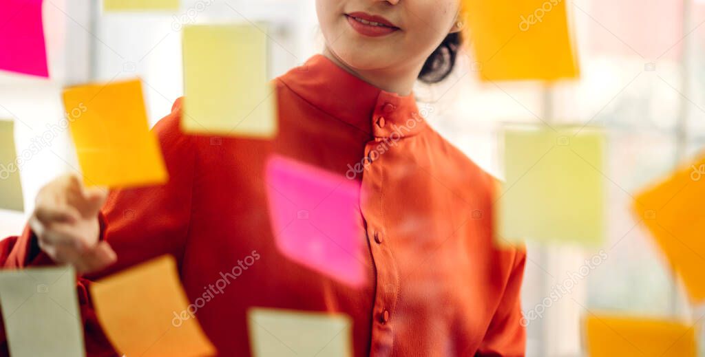 Casual creative happy smile asian business woman planning strategy analysis putting post it stickers note to share idea for startup project on glass board at modern office