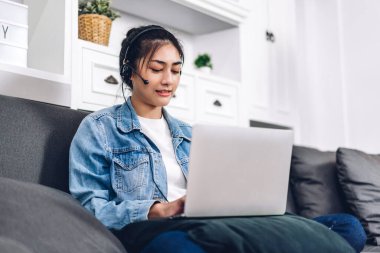 Genç gülümseyen mutlu Asyalı kadın dizüstü bilgisayar kullanarak rahatlıyor ve video konferansında evde sohbet ediyor. Ev konseptinden genç yaratıcı kız kulaklıkla konuşuyor.