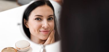 Genç ve güzel bir kadının gülümsemesi taze, sağlıklı bir beyaz tenin aynaya bakışıdır. Yüzüne elle dokunan ve evde krem süren. spa ve güzellik konsepti.